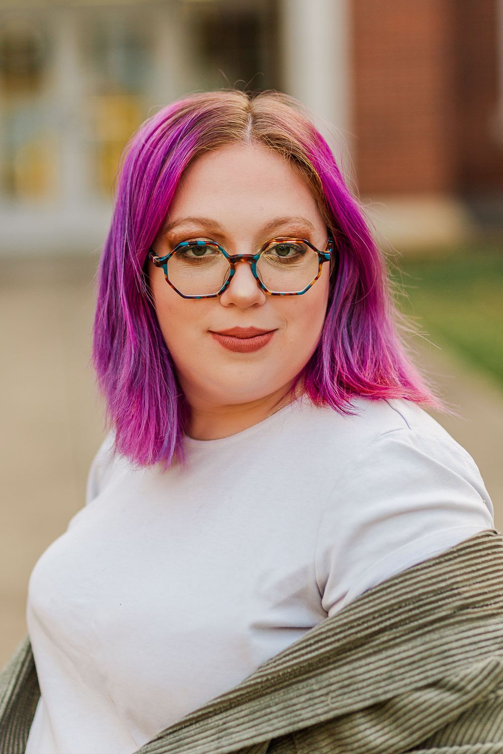Portrait showcasing vibrant pink hair and glasses against red brick building backdrop.