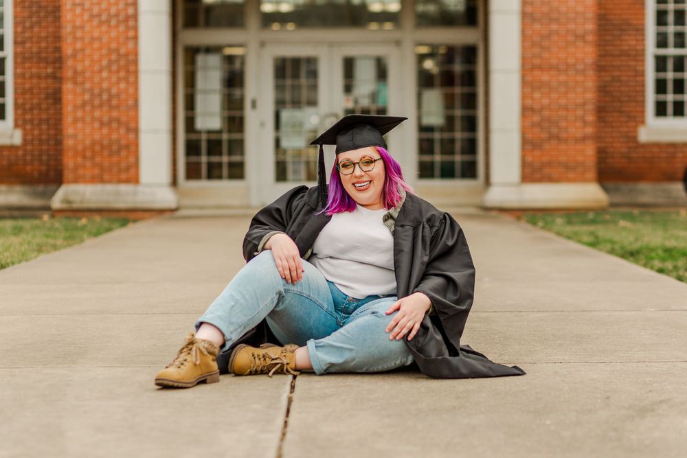 Graduate sitting on sidewalk wearing cap and gown in front of academic building entrance.