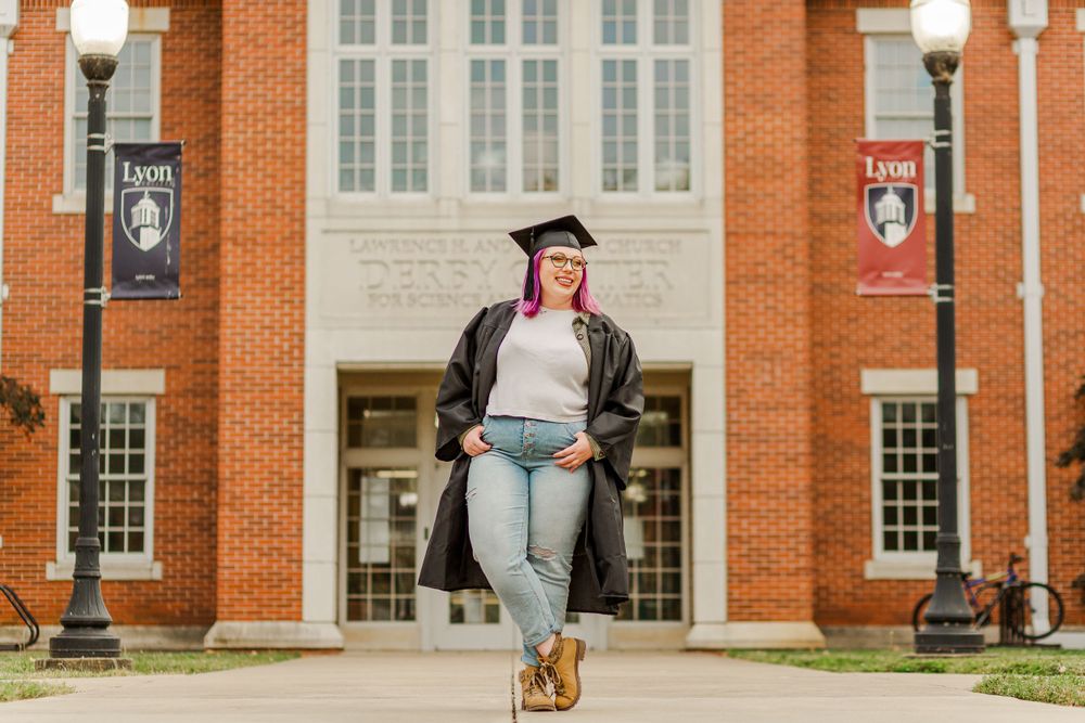 Graduate with pink hair stands in front of brick university building with white columns.