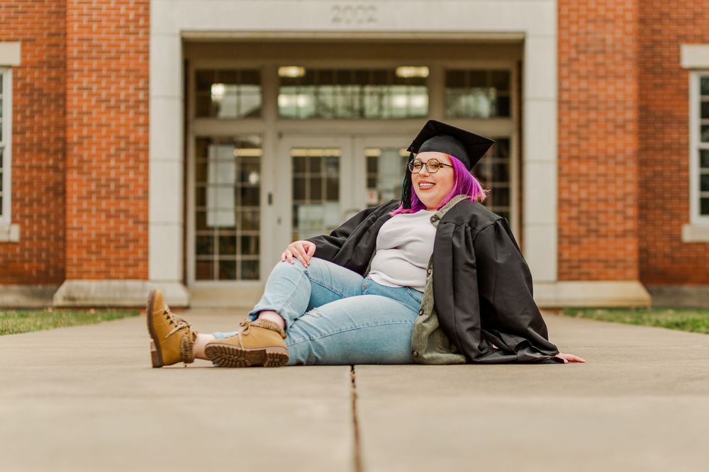 Graduate in academic regalia relaxes on concrete path in front of school building.