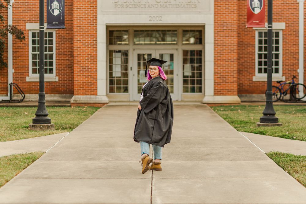 Graduate in black gown poses joyfully on campus walkway in front of brick building.