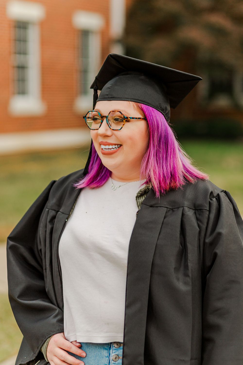 Graduate with vibrant pink hair smiles in academic regalia against brick building.