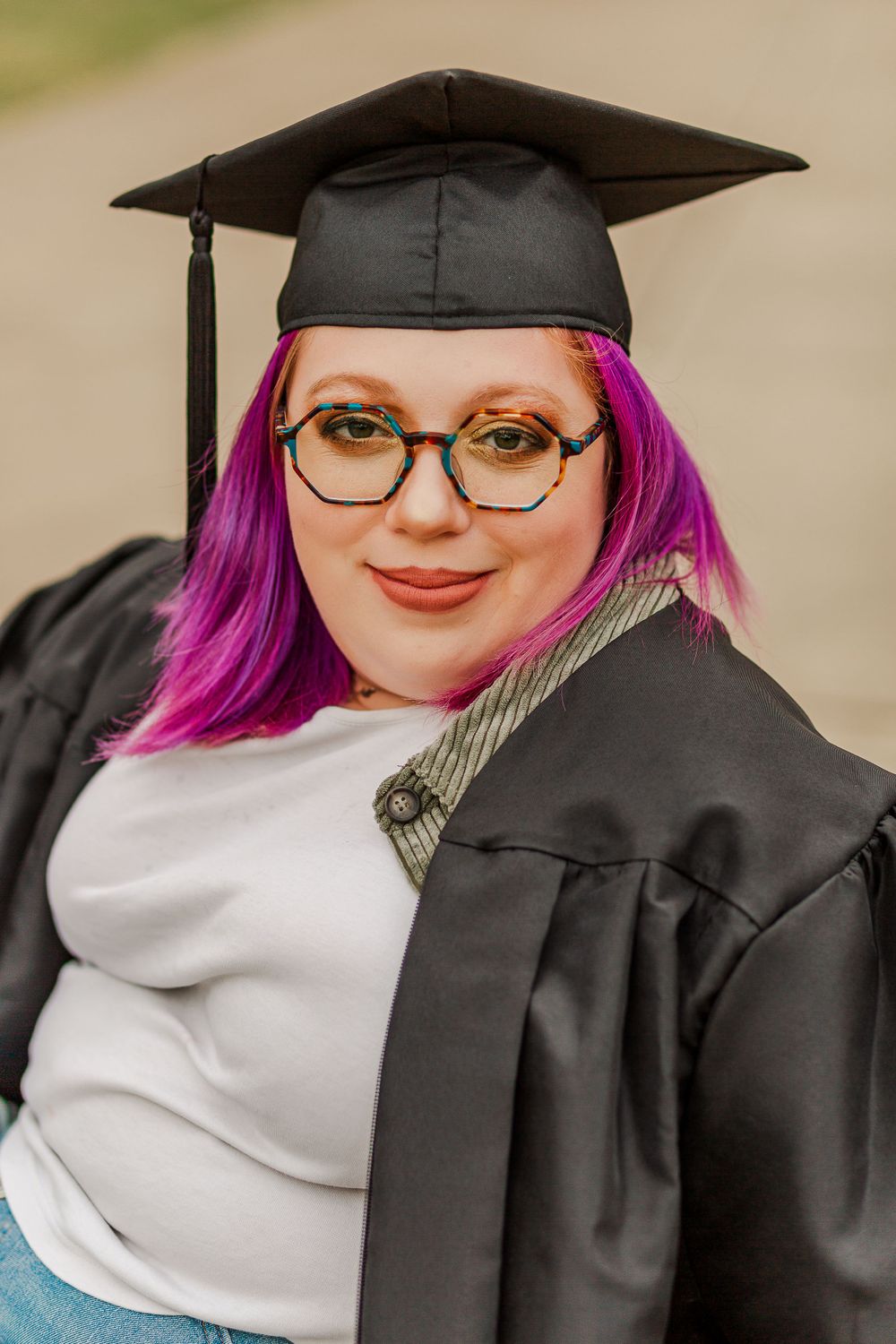 Close-up graduation portrait showing academic cap and bright pink hair against blurred background.