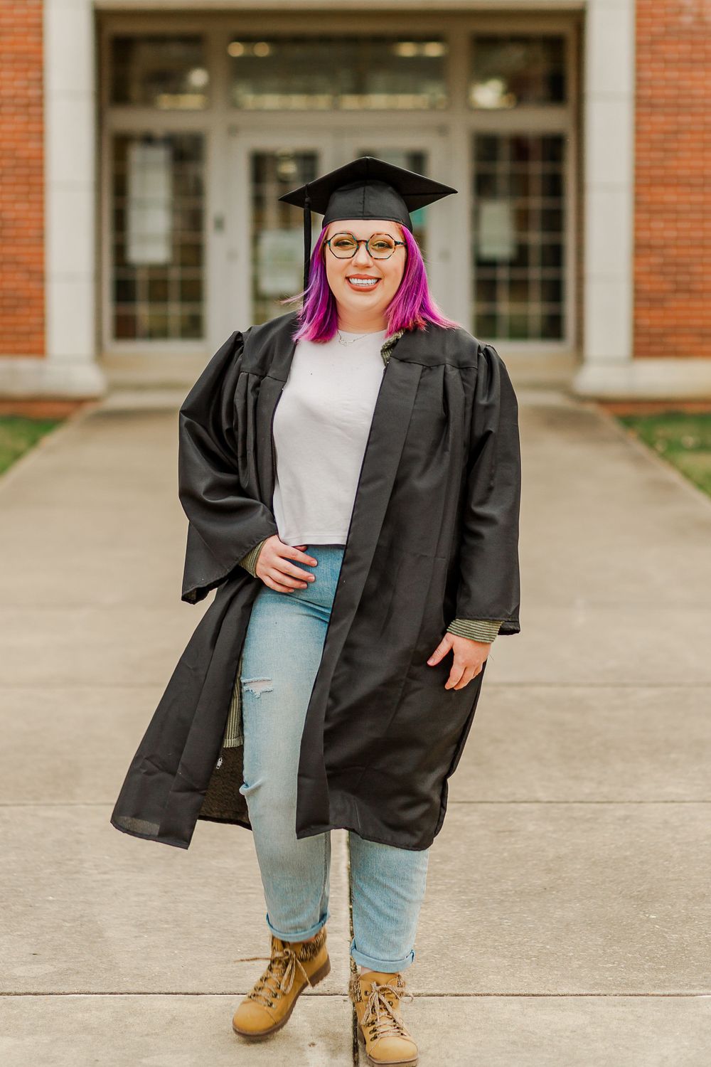Graduate in black cap and gown stands confidently in front of collegiate building entrance.