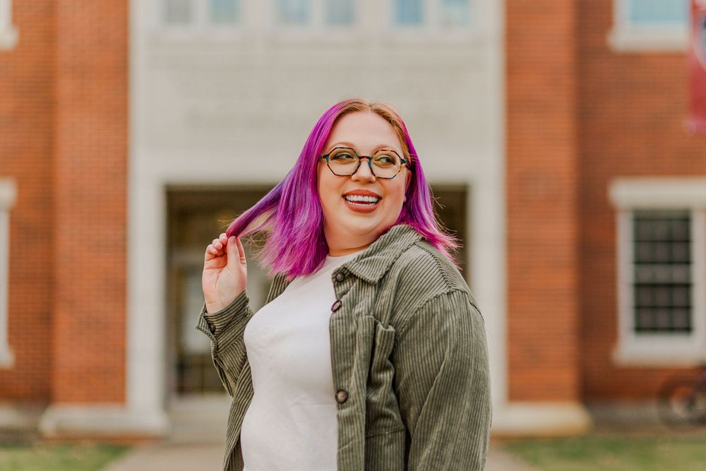 Casual portrait against brick building showing bright pink hair and olive jacket.