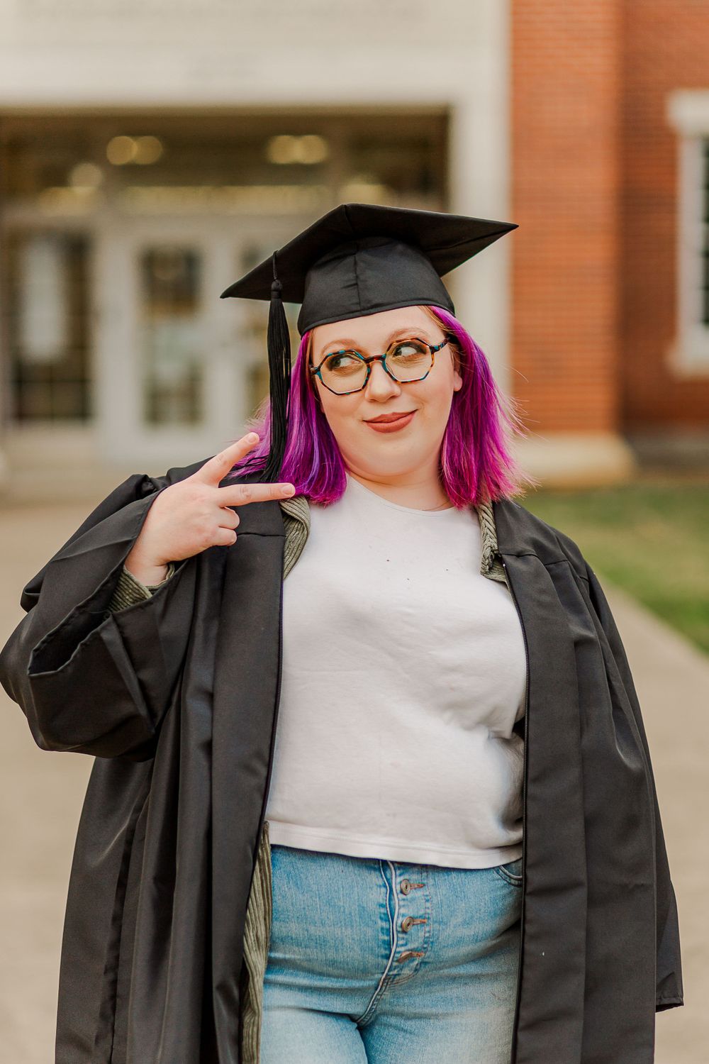Graduate makes peace sign while wearing academic regalia in front of campus architecture.