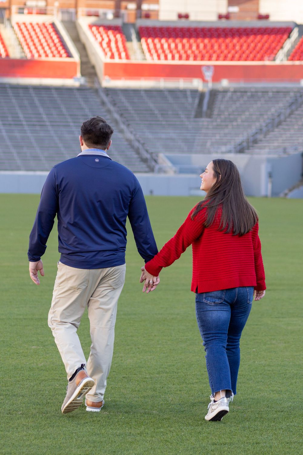 A newly engaged couple walks away holding hands on the NC State football field.