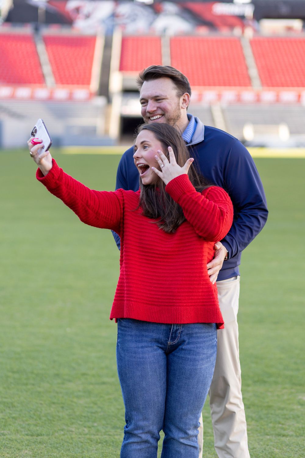 A couple takes a selfie after getting engaged. She is wearing a red sweater and he is in a navy pullover.