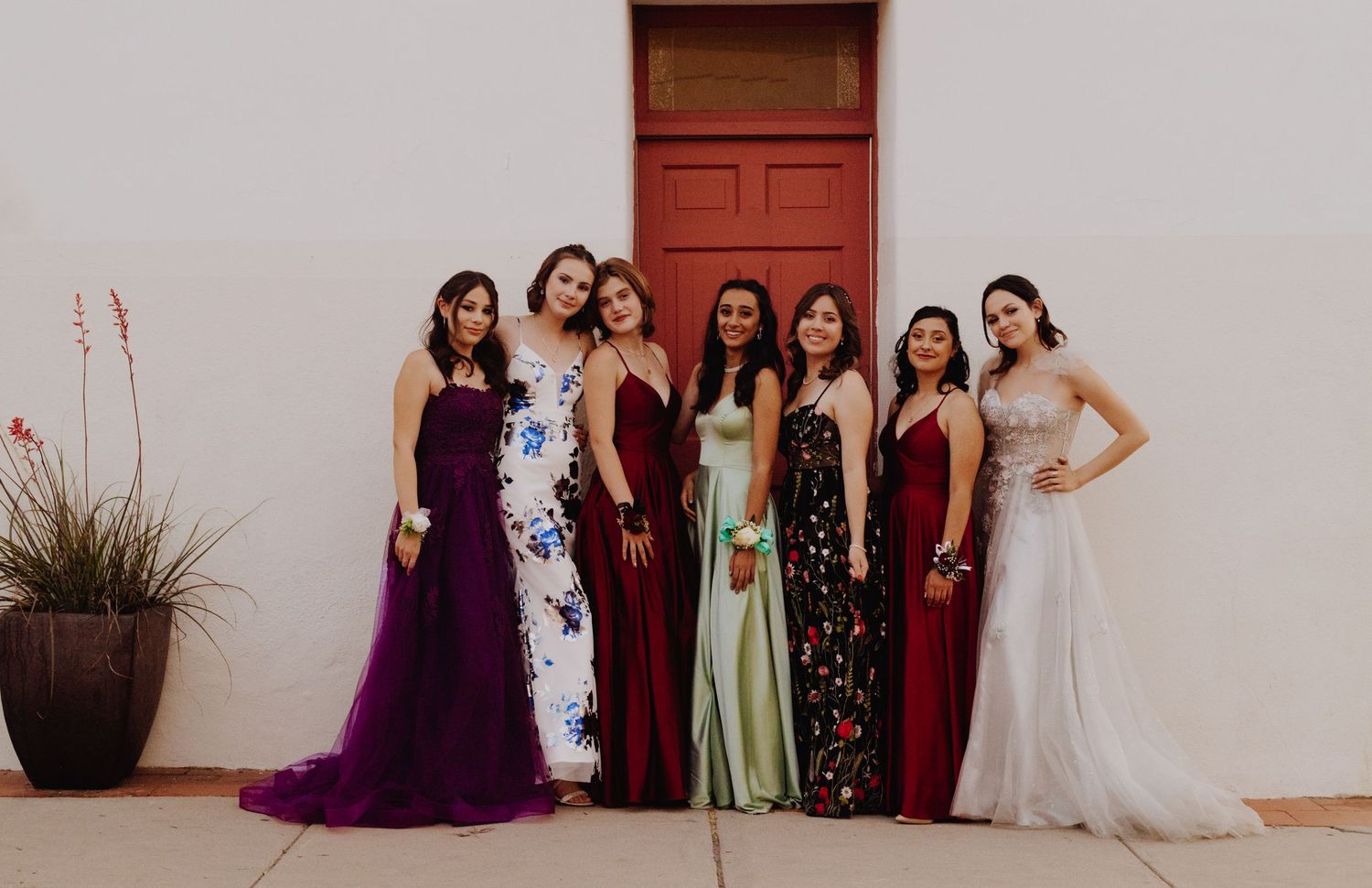 Group photo of wedding party wearing formal dresses against white wall.