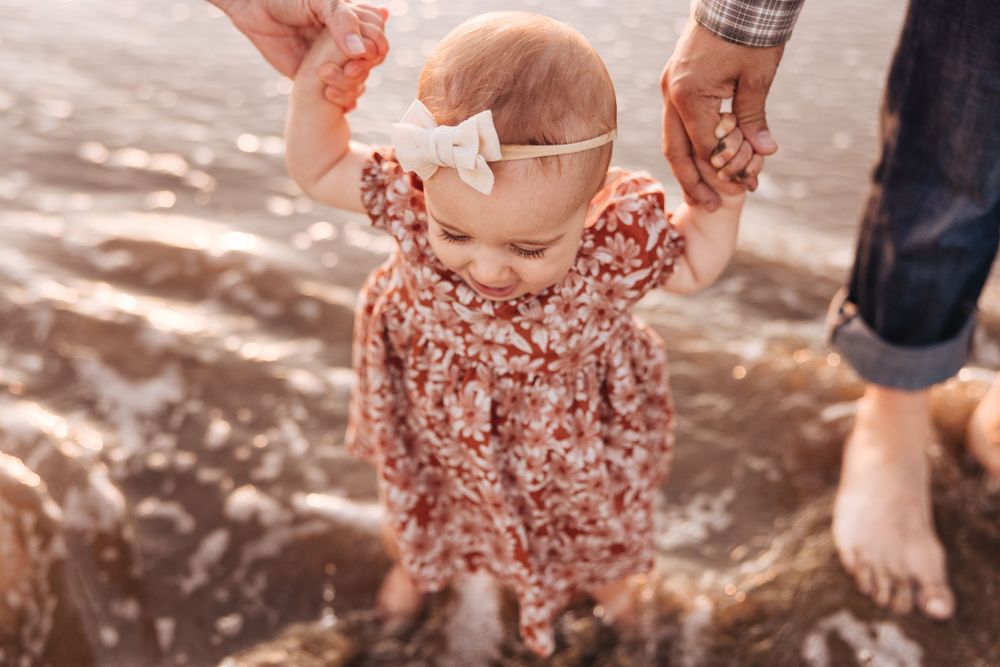 A toddler in a floral dress plays in the ocean waves at sunset.