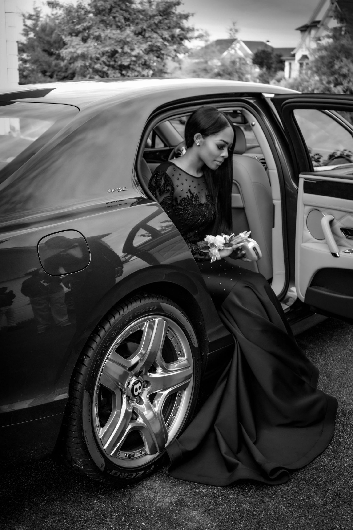 Black and white photo of someone in a formal dress next to a luxury car with custom wheels. Prom photoshoot. Lifestyle photoshoot. L Visions