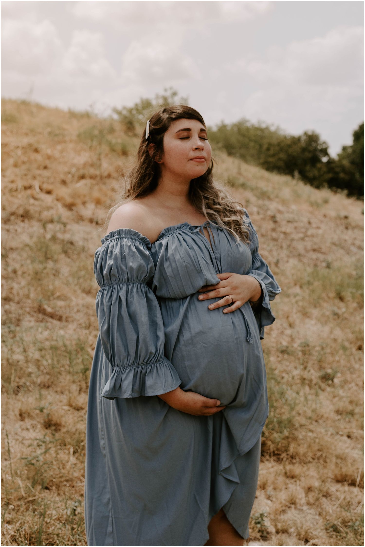 Serene maternity portrait featuring flowing blue off-shoulder dress in natural outdoor setting.