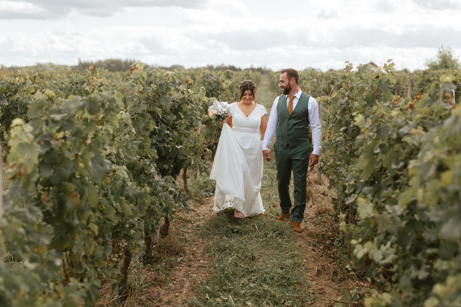 A romantic wedding couple walks hand in hand through rows of grapevines at a scenic vineyard.