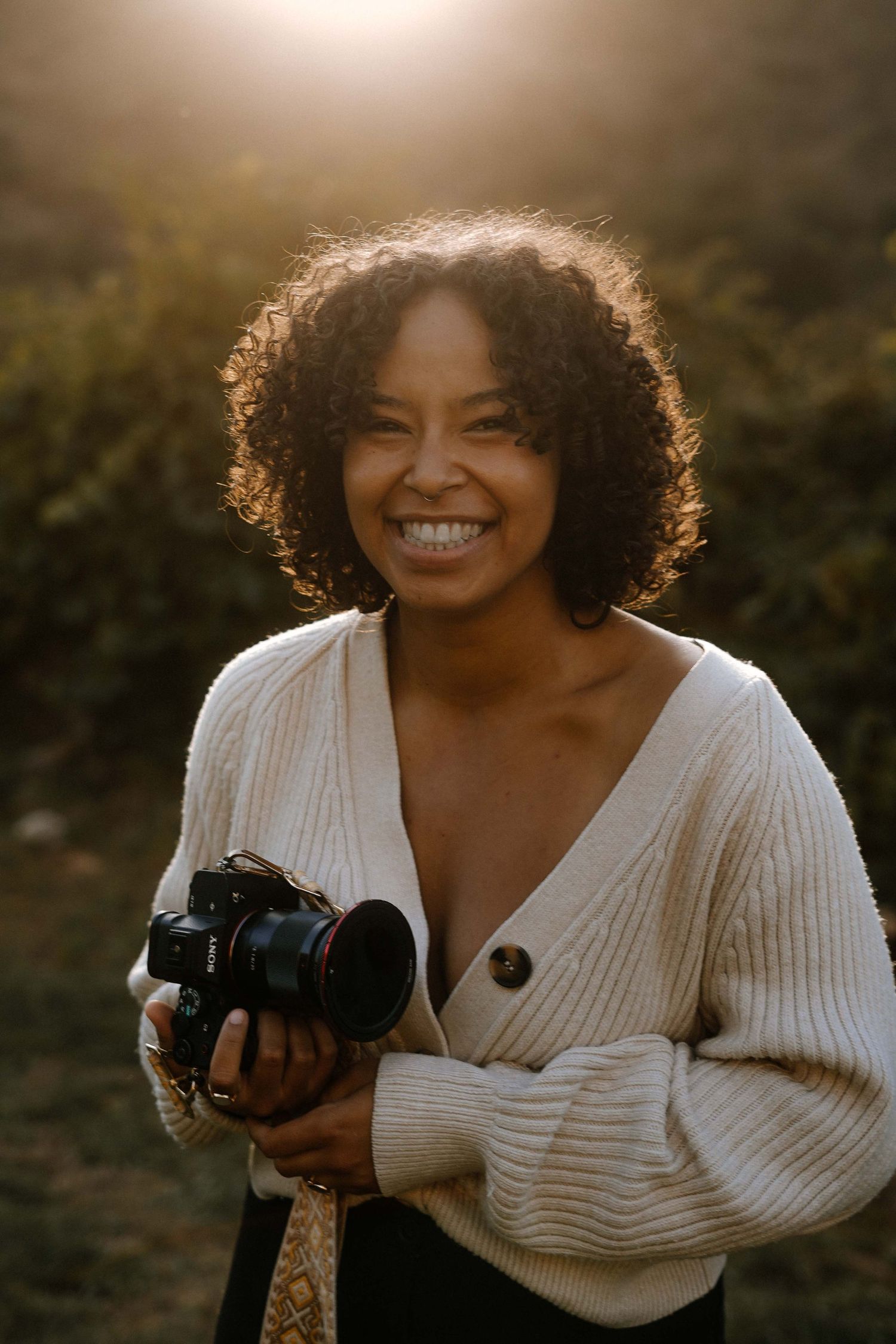 Professional photographer in a white cardigan sweater holding a camera during golden hour lighting.