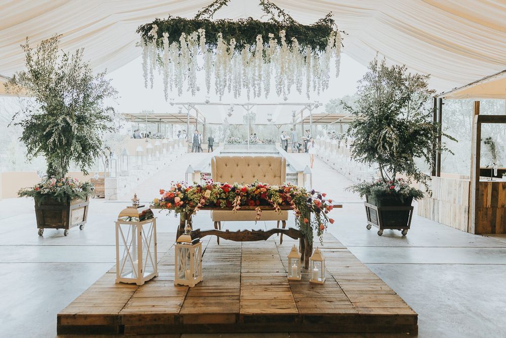Modern white tent reception space with wooden head table and hanging greenery installations.