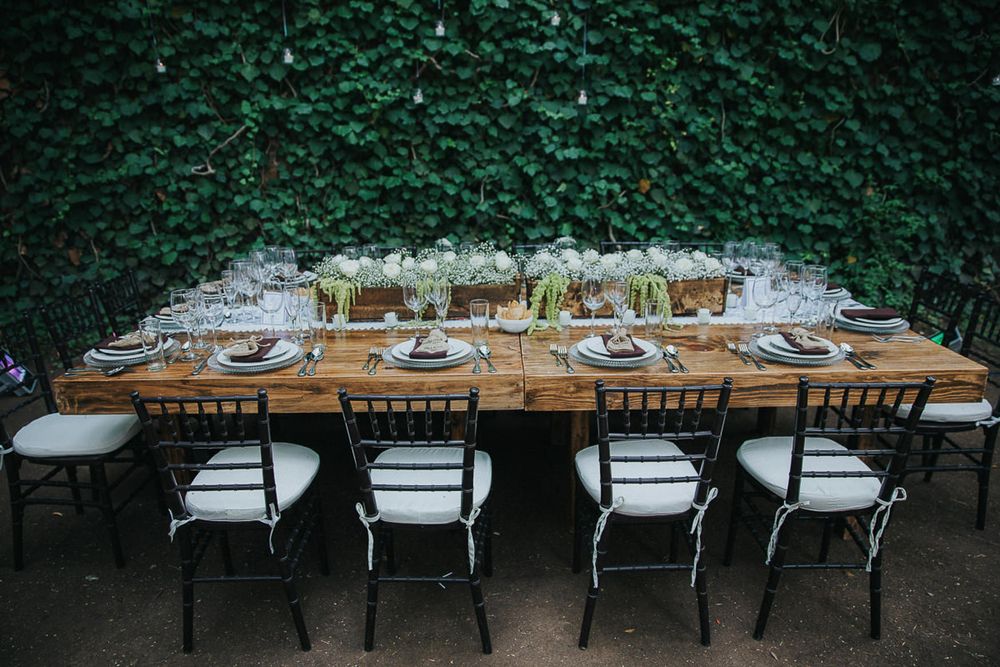 Long farmhouse table with place settings against a green hedge wall for an intimate outdoor dinner.