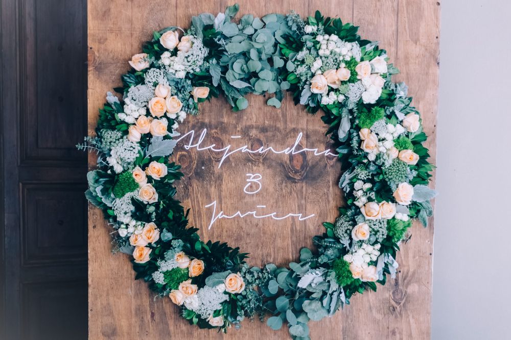 A rustic wooden backdrop displays an elegant green and white floral wreath with a handwritten text in the center.