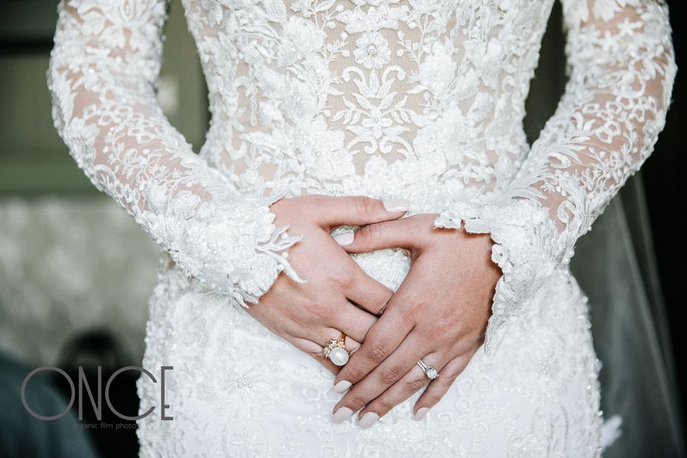 Detail shot of intricate lace wedding dress with hands showing elegant engagement and wedding rings.