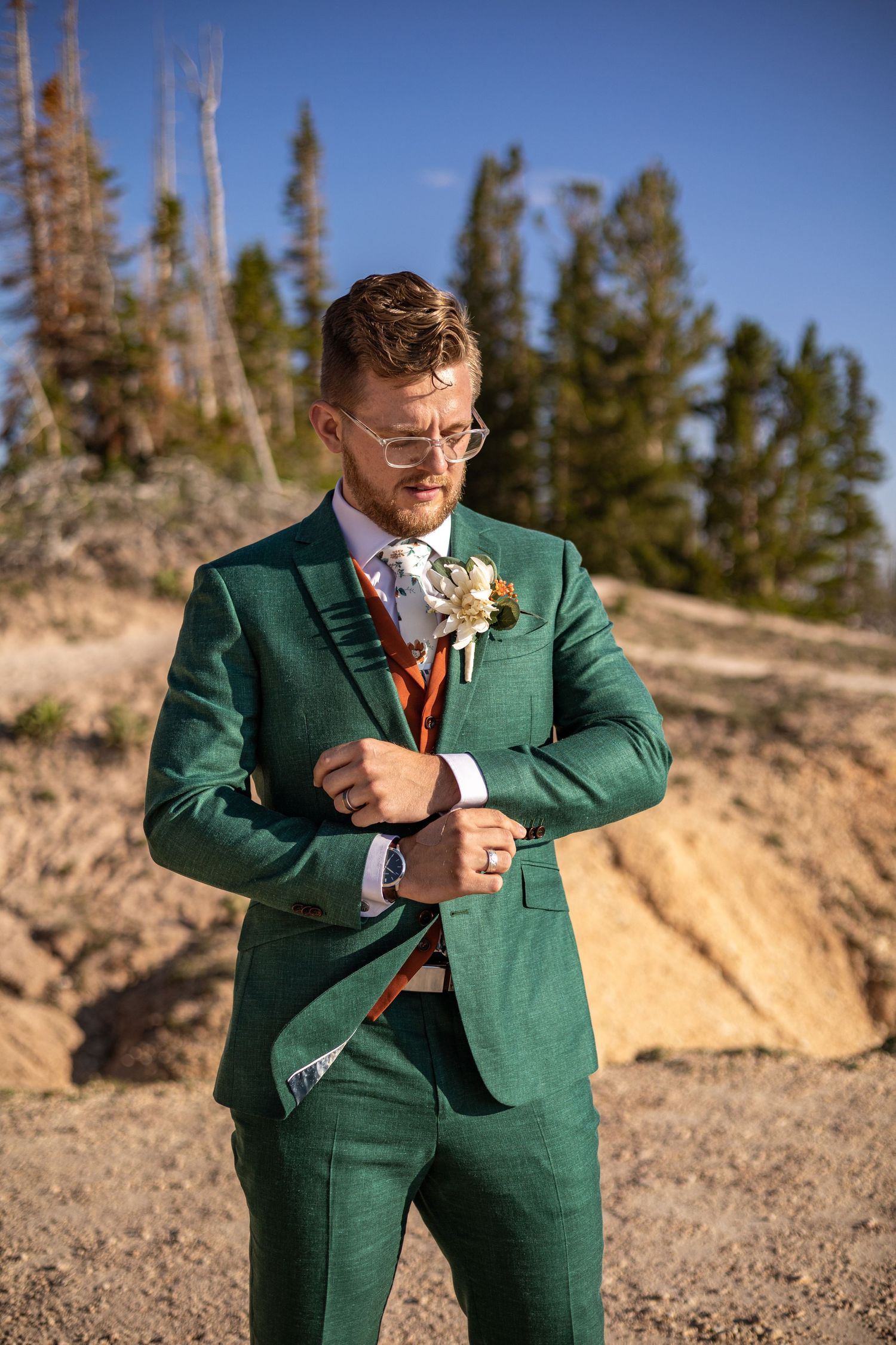 Groom in emerald green suit adjusts cufflinks against mountain landscape.