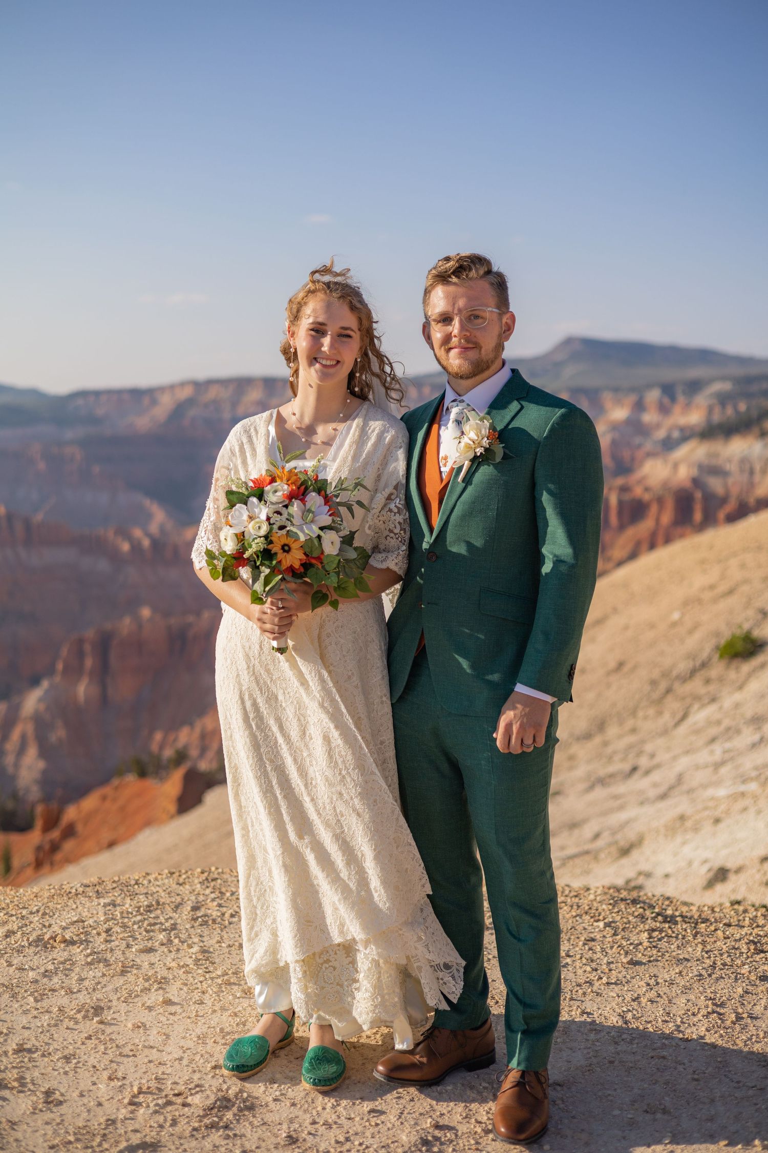 Newlyweds in green suit and lace dress pose together on a scenic mountain overlook.