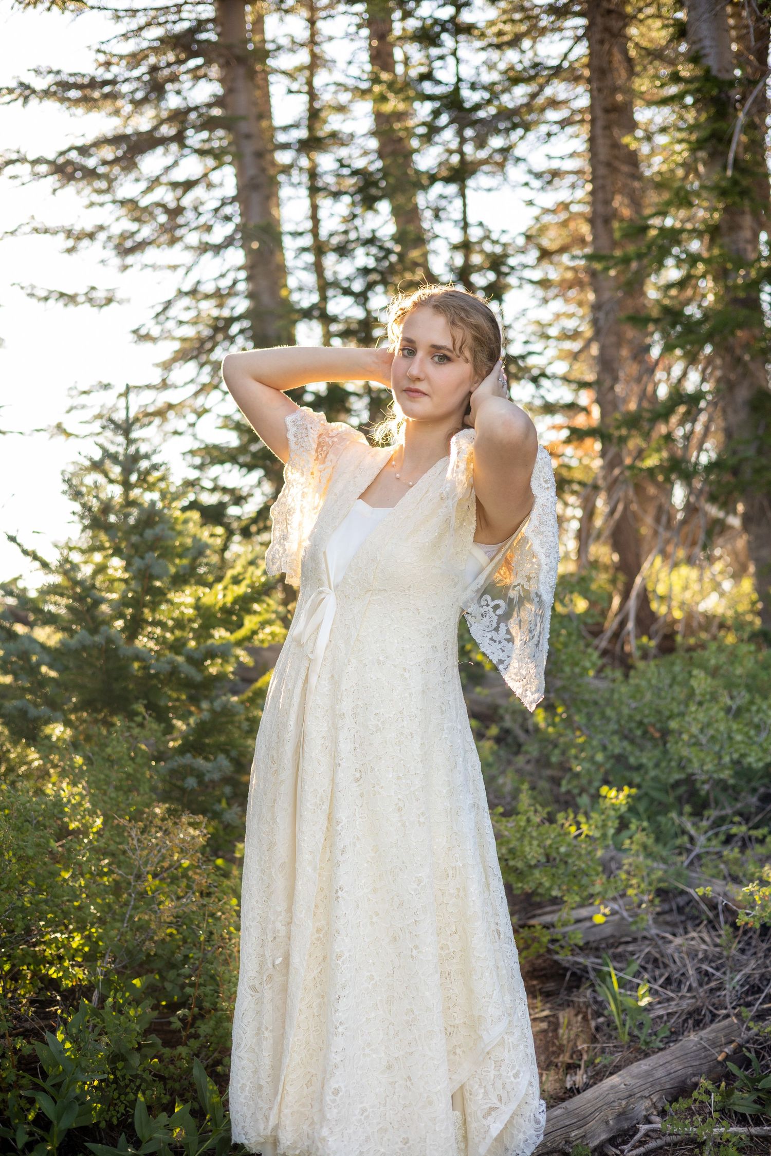 Bride in lace dress poses playfully in a sunlit forest setting.