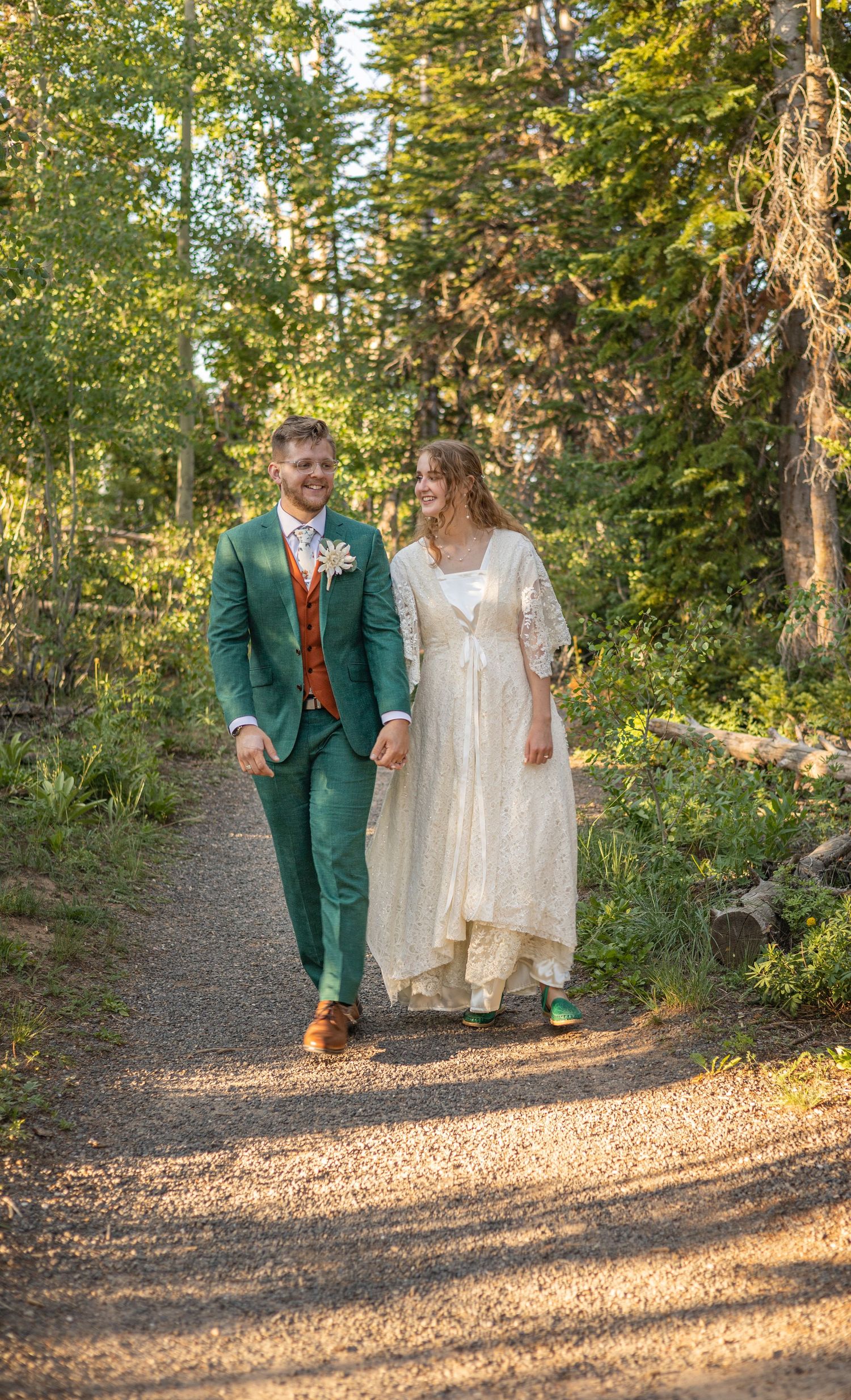 A couple walks hand in hand along a forest path during golden hour.