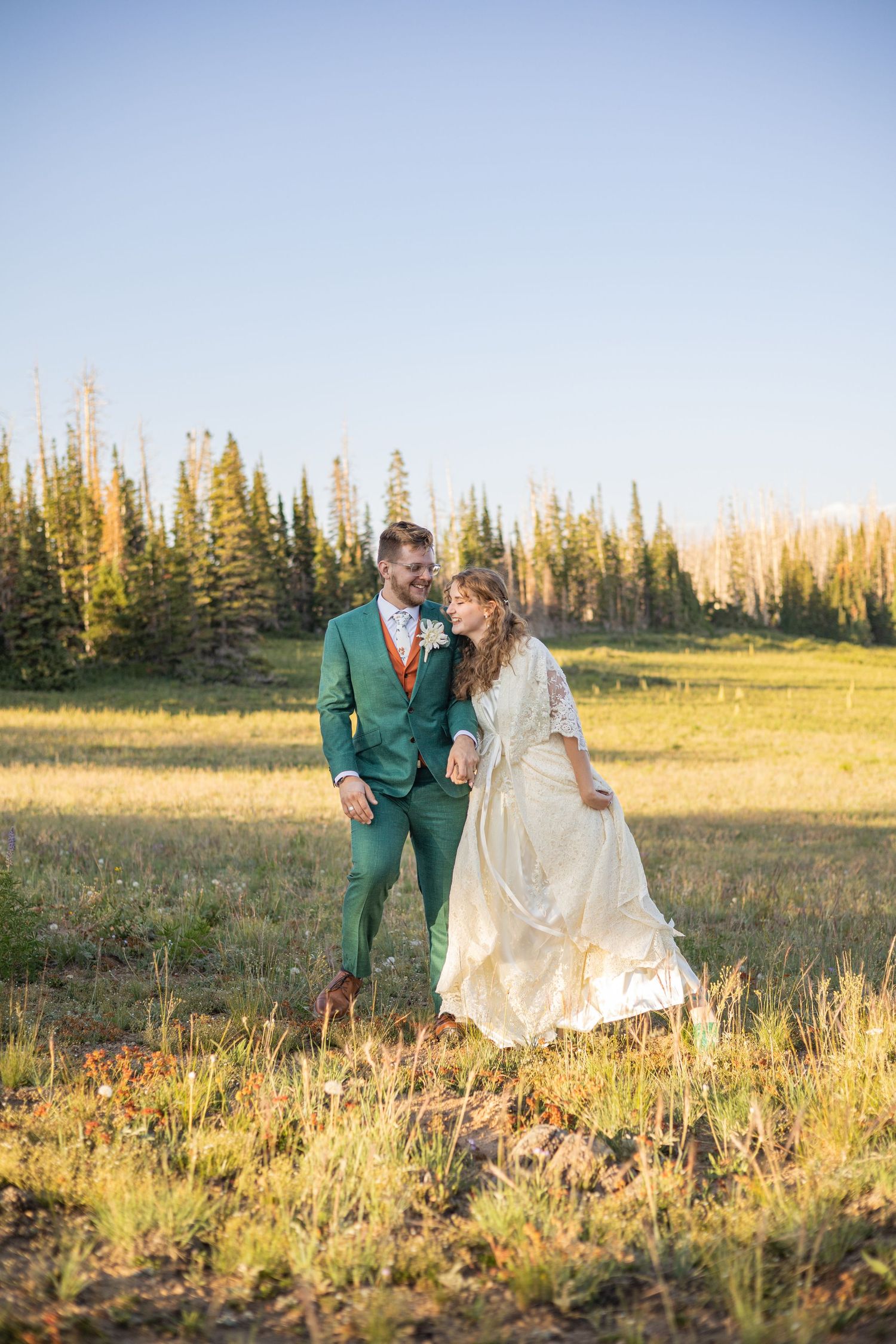 A couple in wedding attire embraces in a sunlit meadow with evergreen trees in the background.