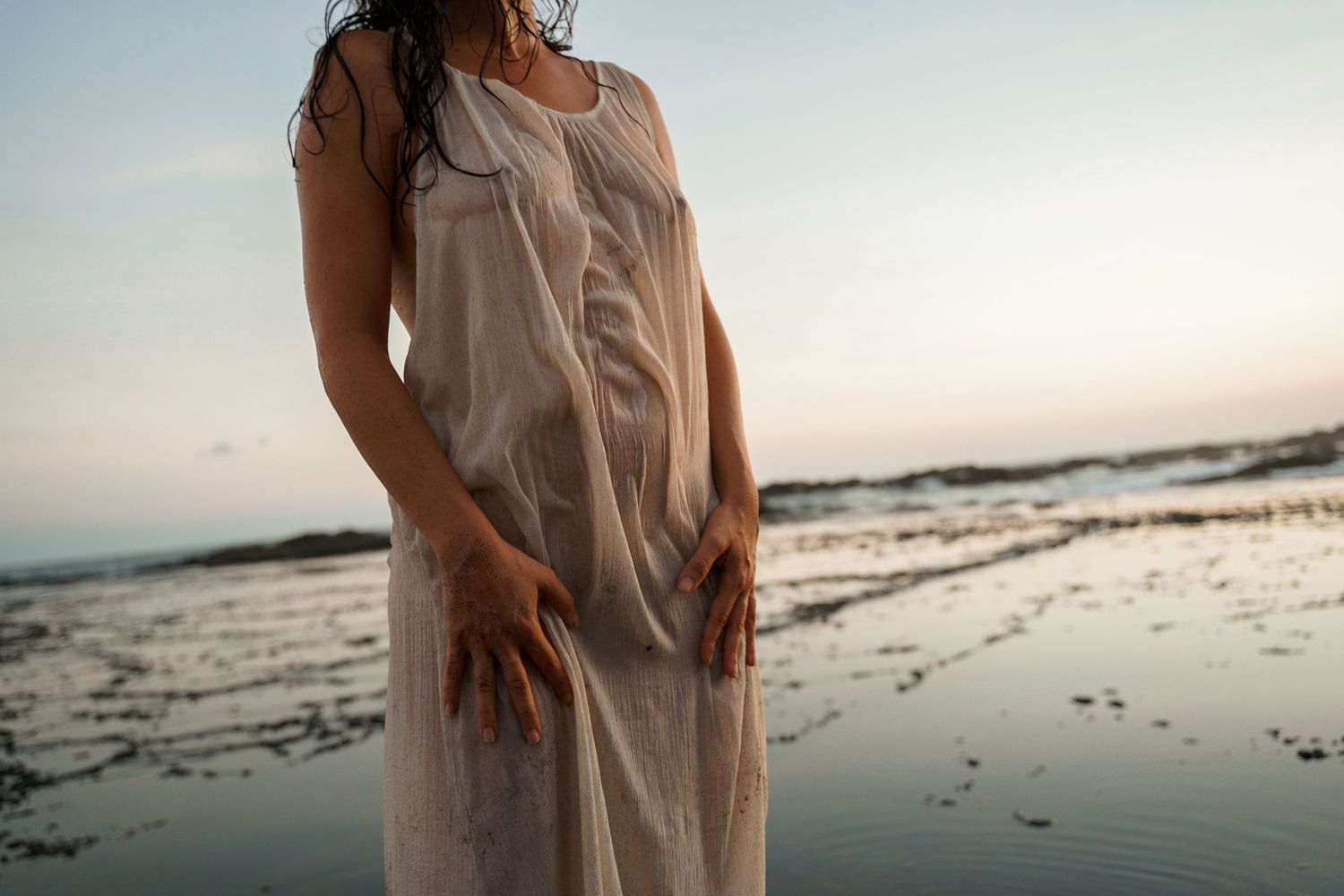 Silhouetted figure in flowing white dress stands on beach at dusk.