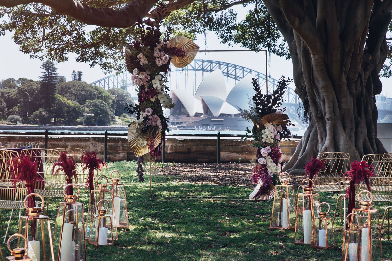 Elegant outdoor wedding aisle with floral arch, candles, and Sydney Opera House view.