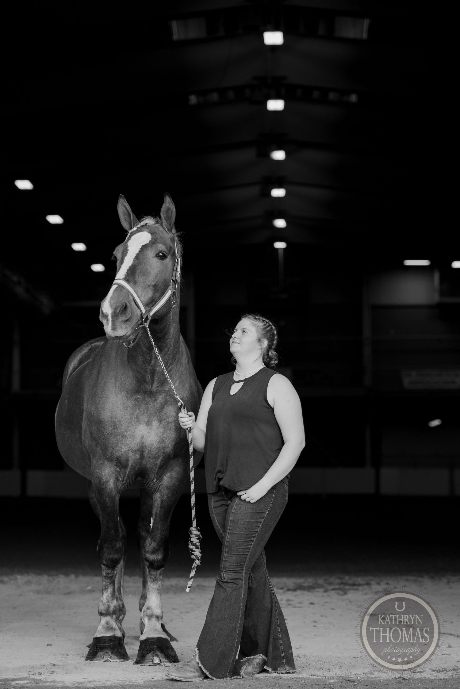 A black and white portrait of a horse and handler standing in an indoor equestrian arena.
