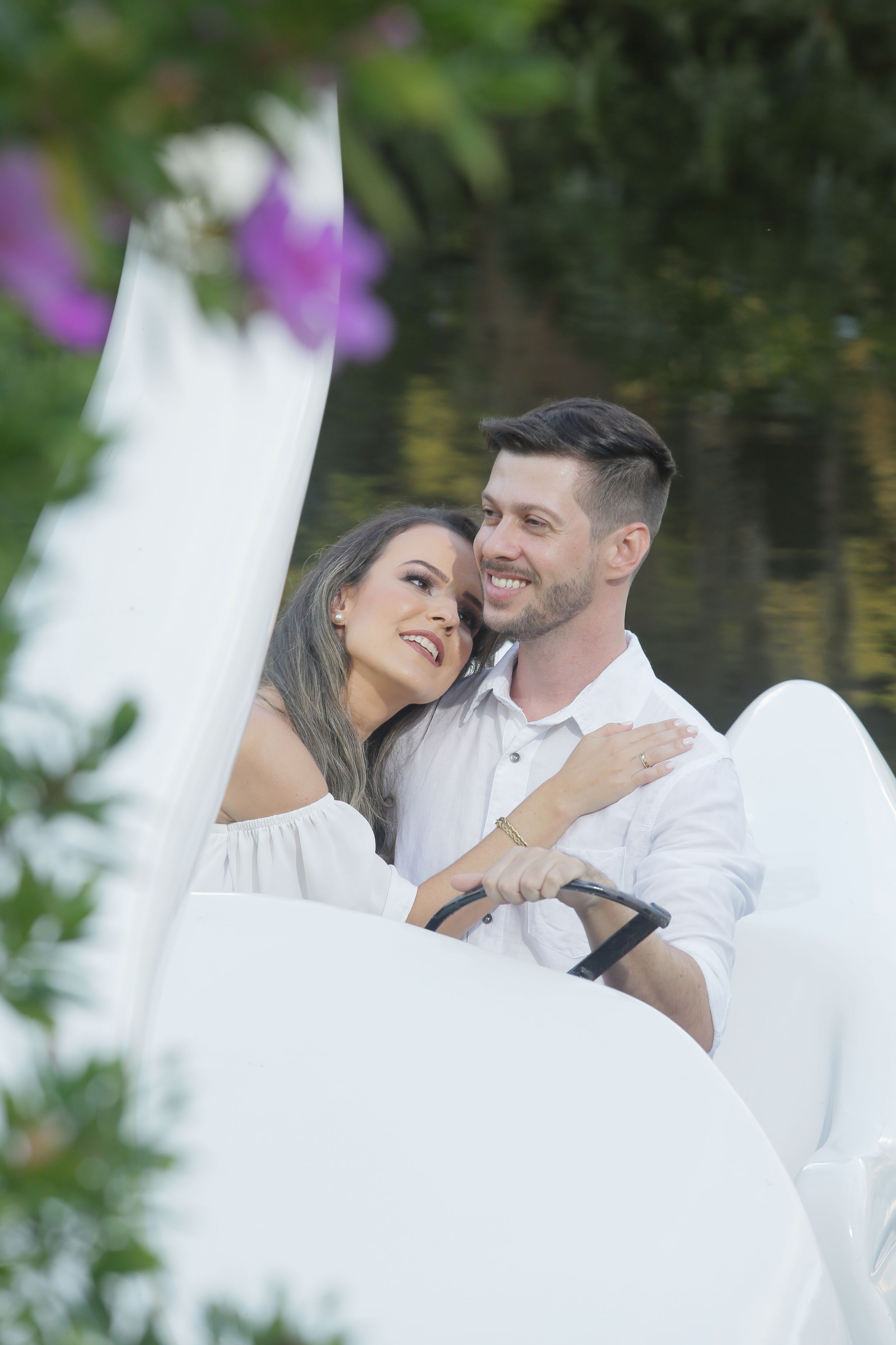 Couple shares romantic moment on swan boat with purple flowers in background.