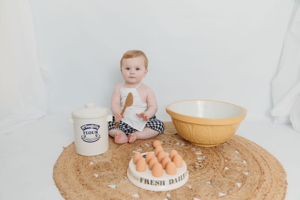 Baby sitting on round jute rug with baking ingredients and mixing bowl.