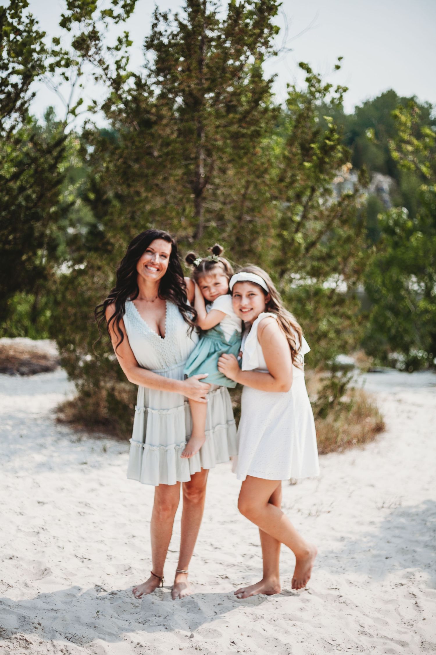 Three people in light summer dresses pose together on a sandy beach near pine trees.