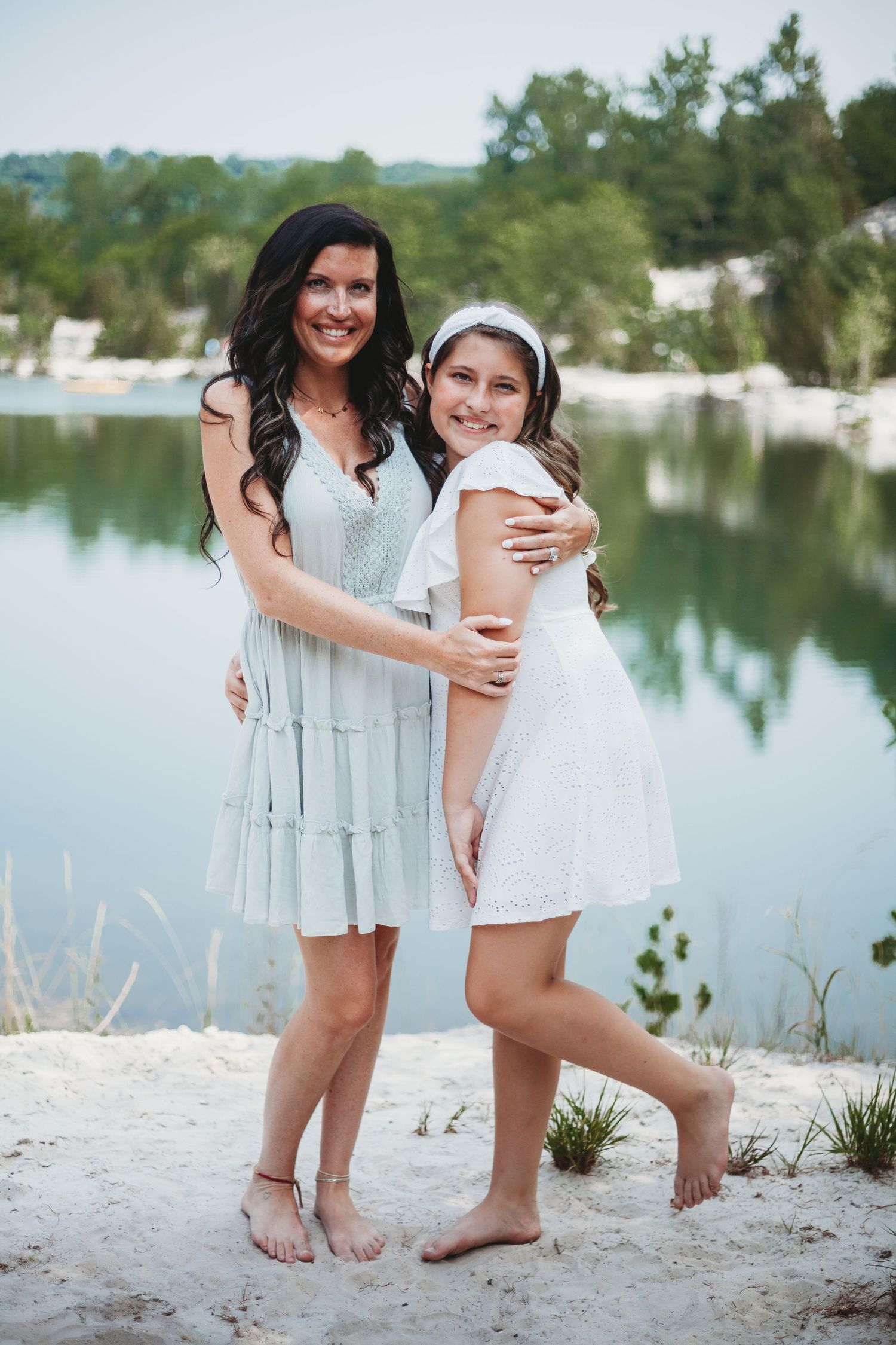 Two people in light dresses embrace while standing on a sandy beach near a lake.