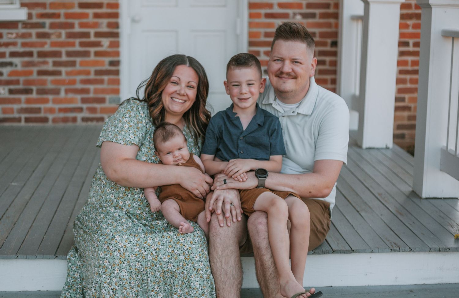 A family sits together on wooden porch steps against a brick and white backdrop.
