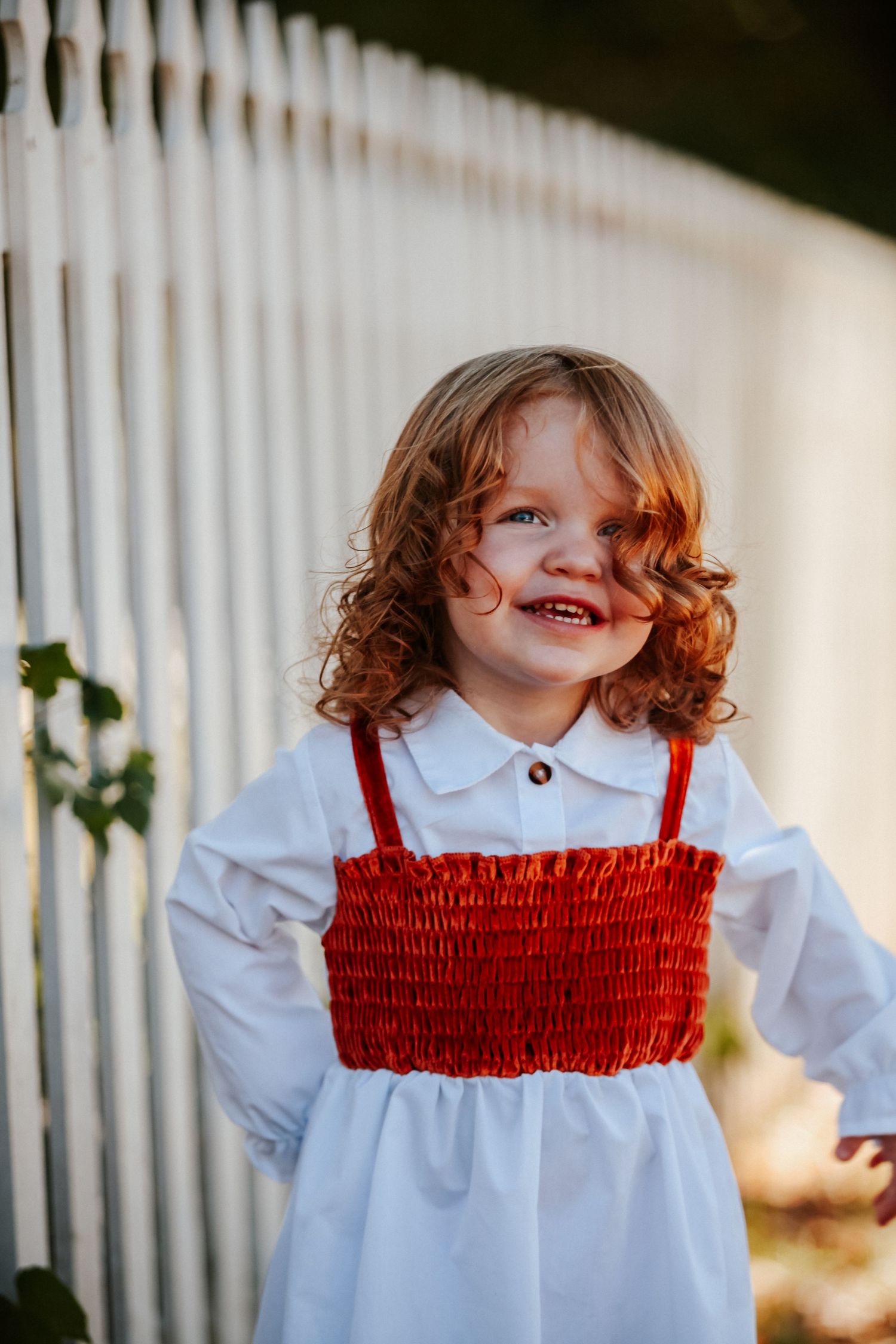 Young child in white dress with red smocked top smiling against white picket fence.