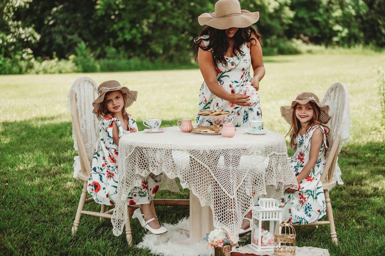 Outdoor tea party scene with floral dresses and lace tablecloth in a garden setting.