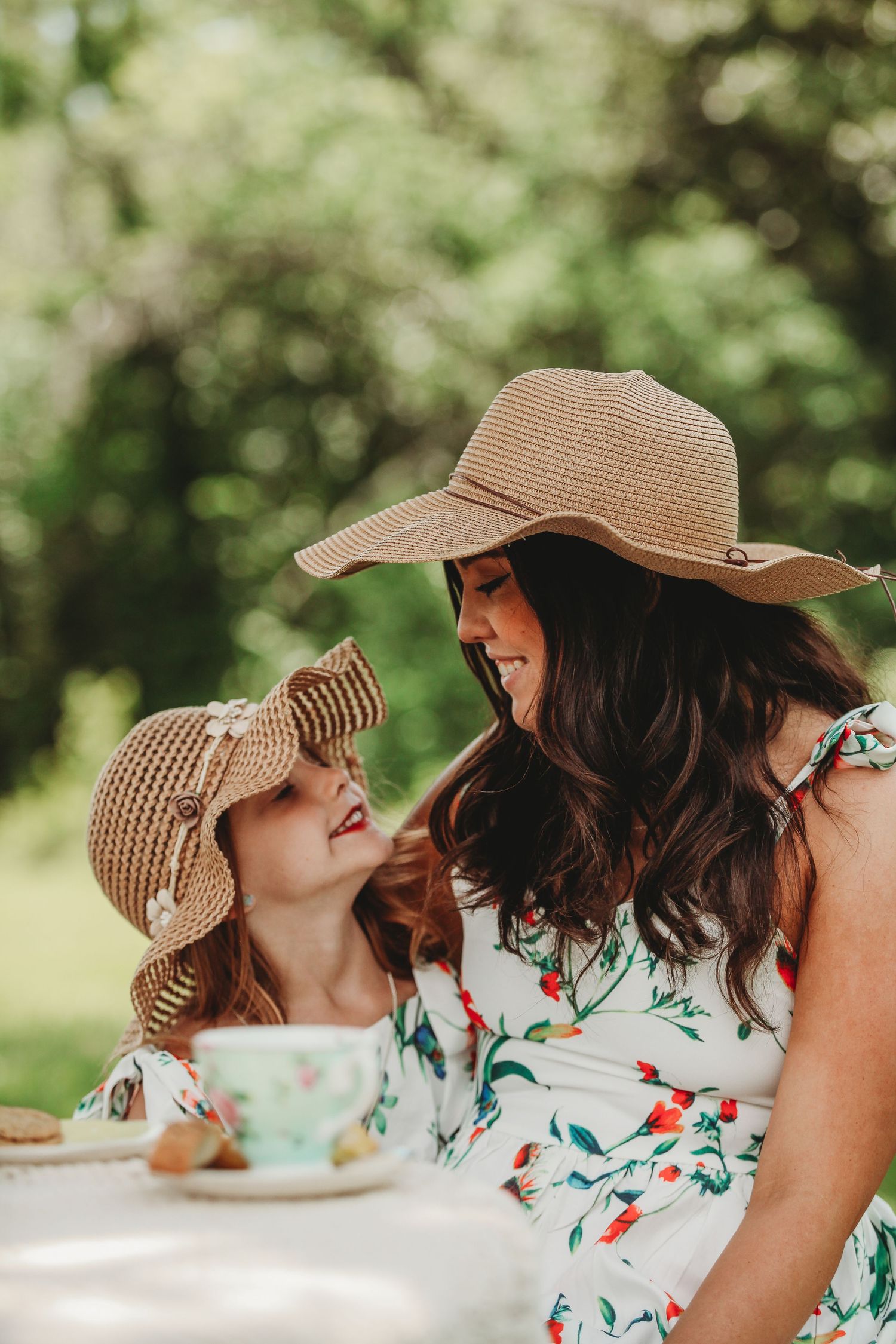 Two people in matching floral dresses and straw hats share a joyful moment over tea outdoors.