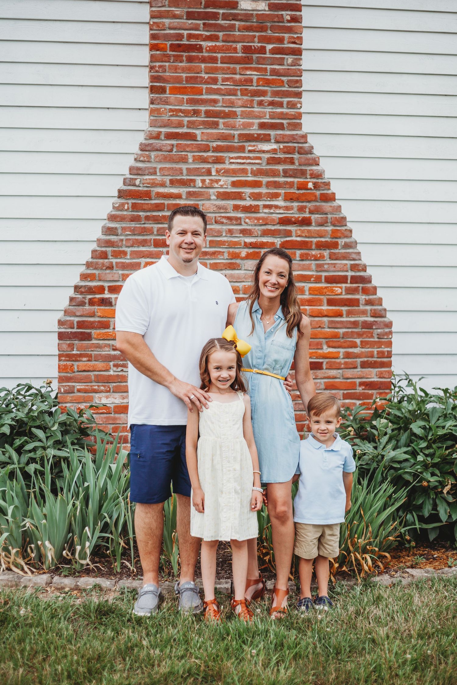 A family stands together against a white house with red brick chimney surrounded by garden plants.