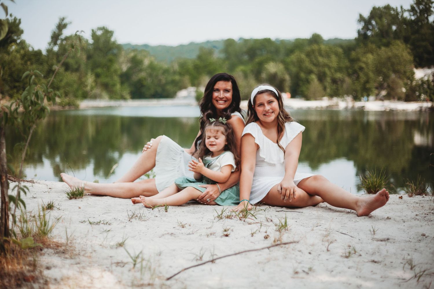 Three people relaxing on white sand beside a peaceful lake.