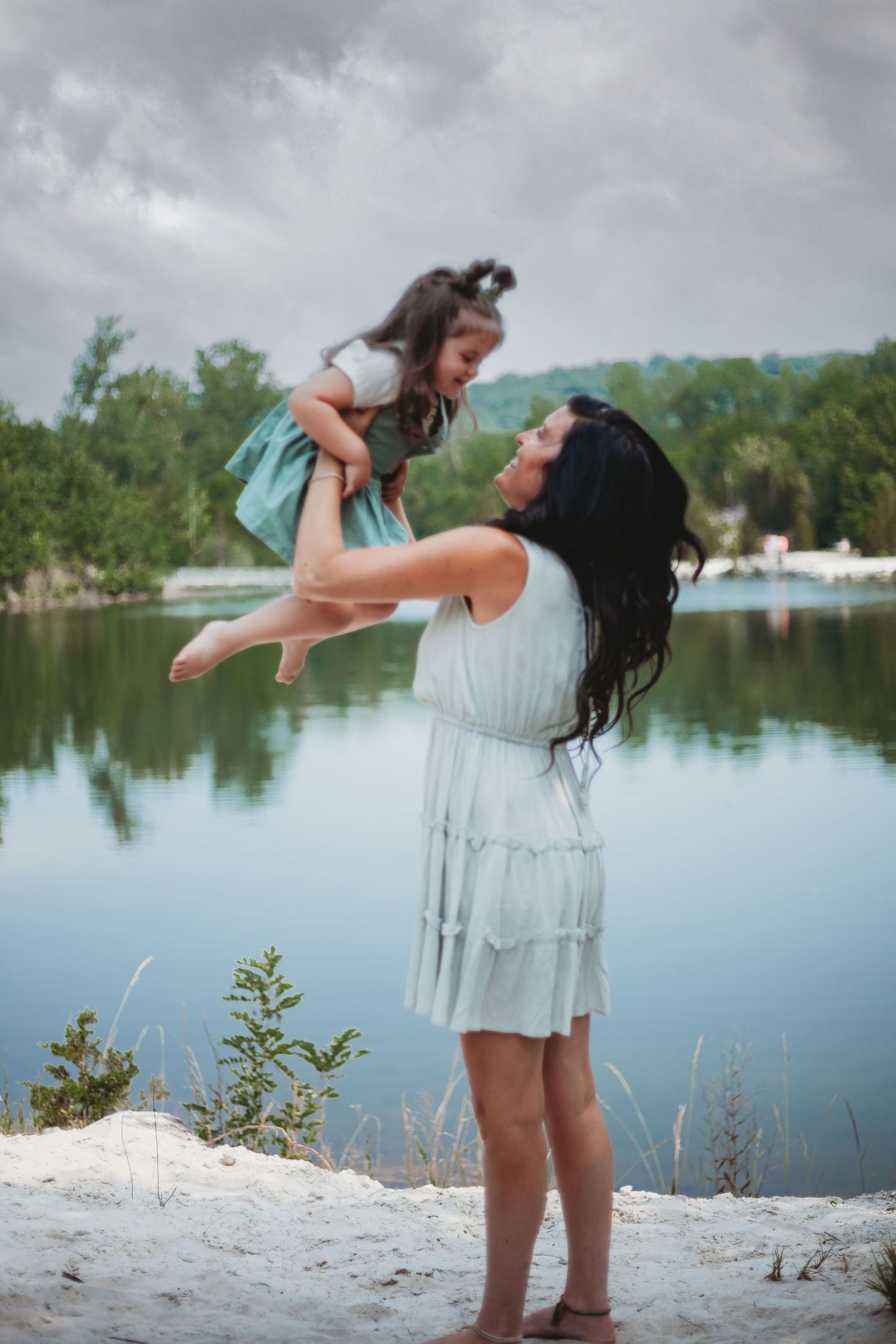A tender moment captured by a lake as a woman in a light dress lifts a small child against a serene water backdrop.