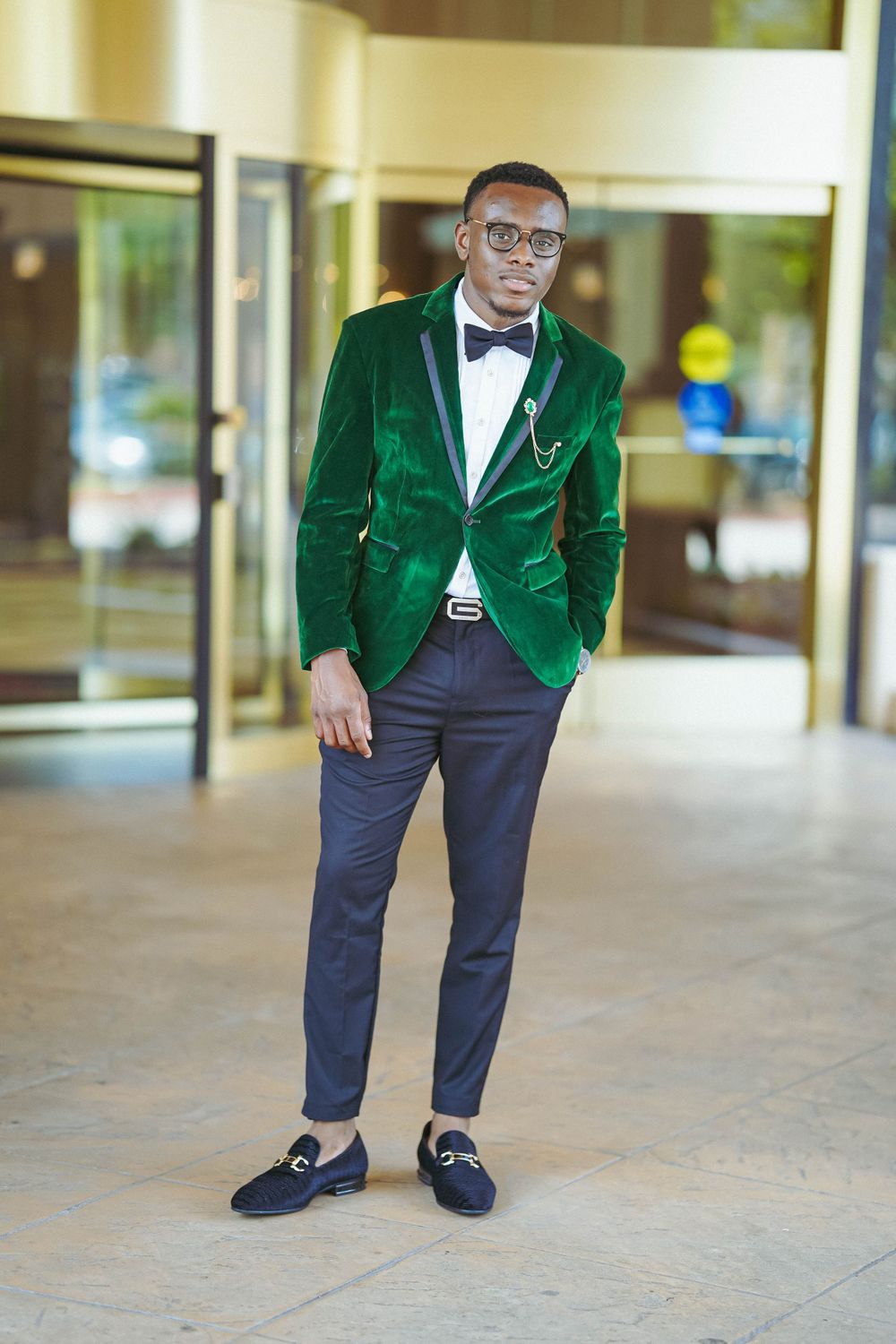Fashionable man wearing emerald green velvet blazer and bowtie stands in upscale venue.