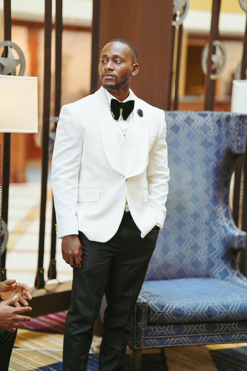 Elegant man in white dinner jacket poses next to blue upholstered chair in formal setting.
