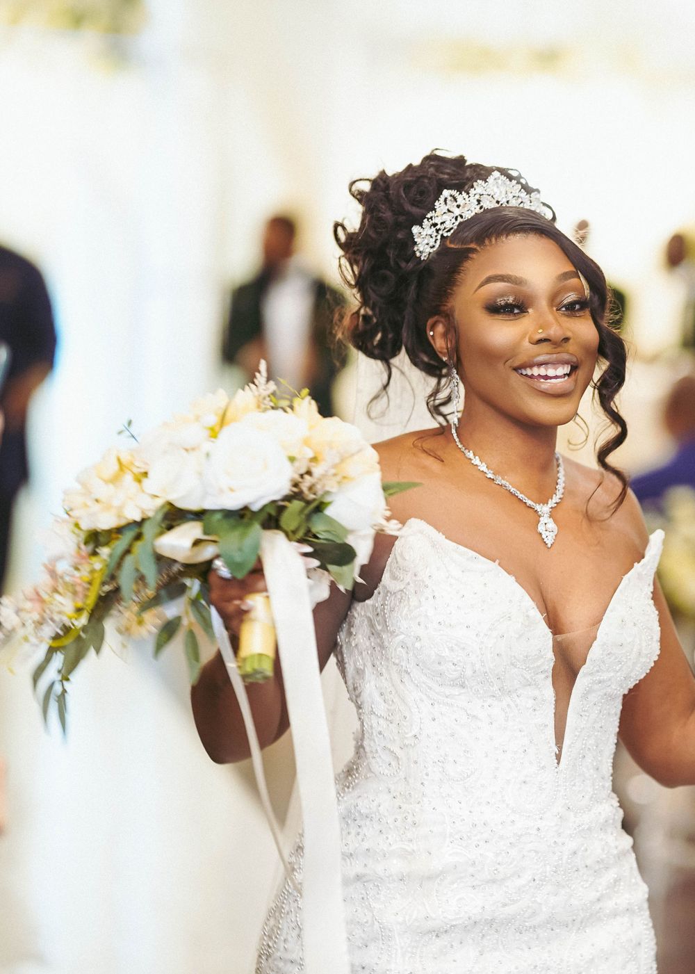 A radiant bride wears a fitted lace gown with plunging neckline and sparkly tiara while holding a white bouquet.