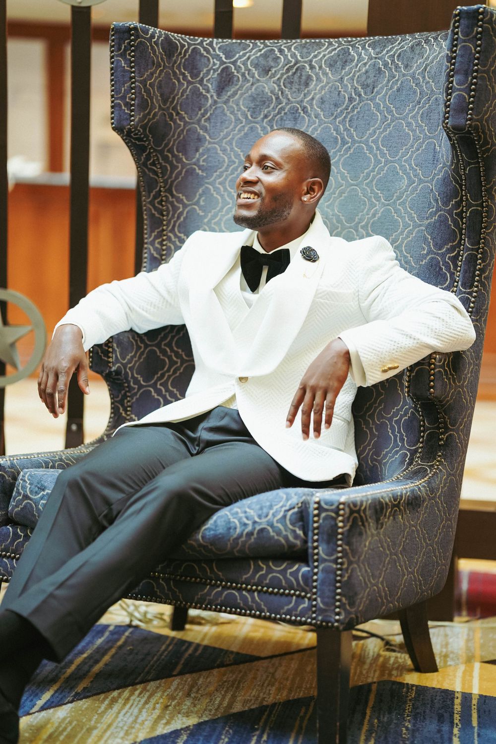 Relaxed groom reclines on patterned chair wearing classic white tuxedo and black bowtie.