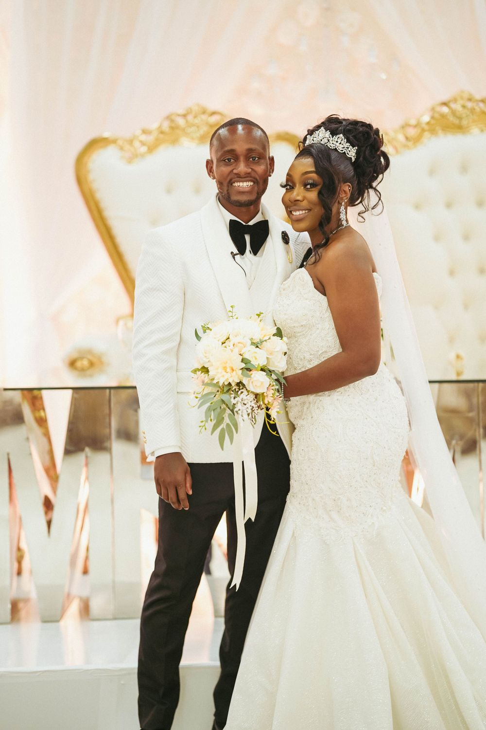Newlyweds pose together against ornate backdrop, groom in white tuxedo and bride in lace mermaid gown.