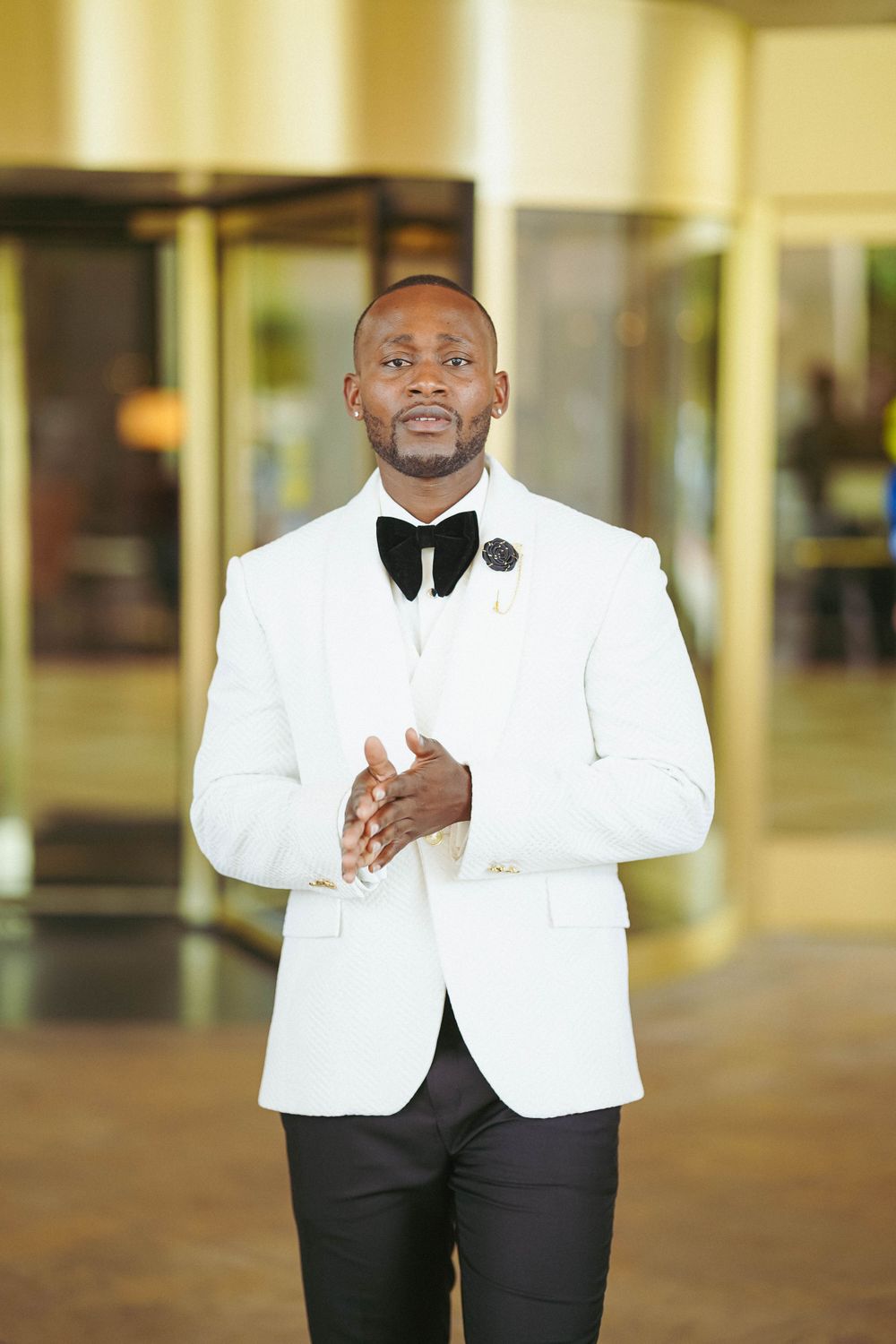 Dapper groom in white dinner jacket and bowtie smiles confidently while standing in a formal venue.