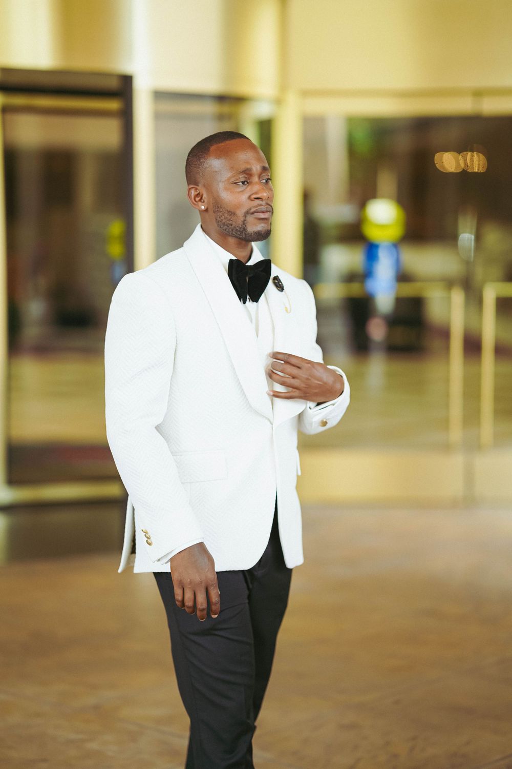 Stylish groom poses in a classic white tuxedo jacket and black pants in an elegant indoor setting.