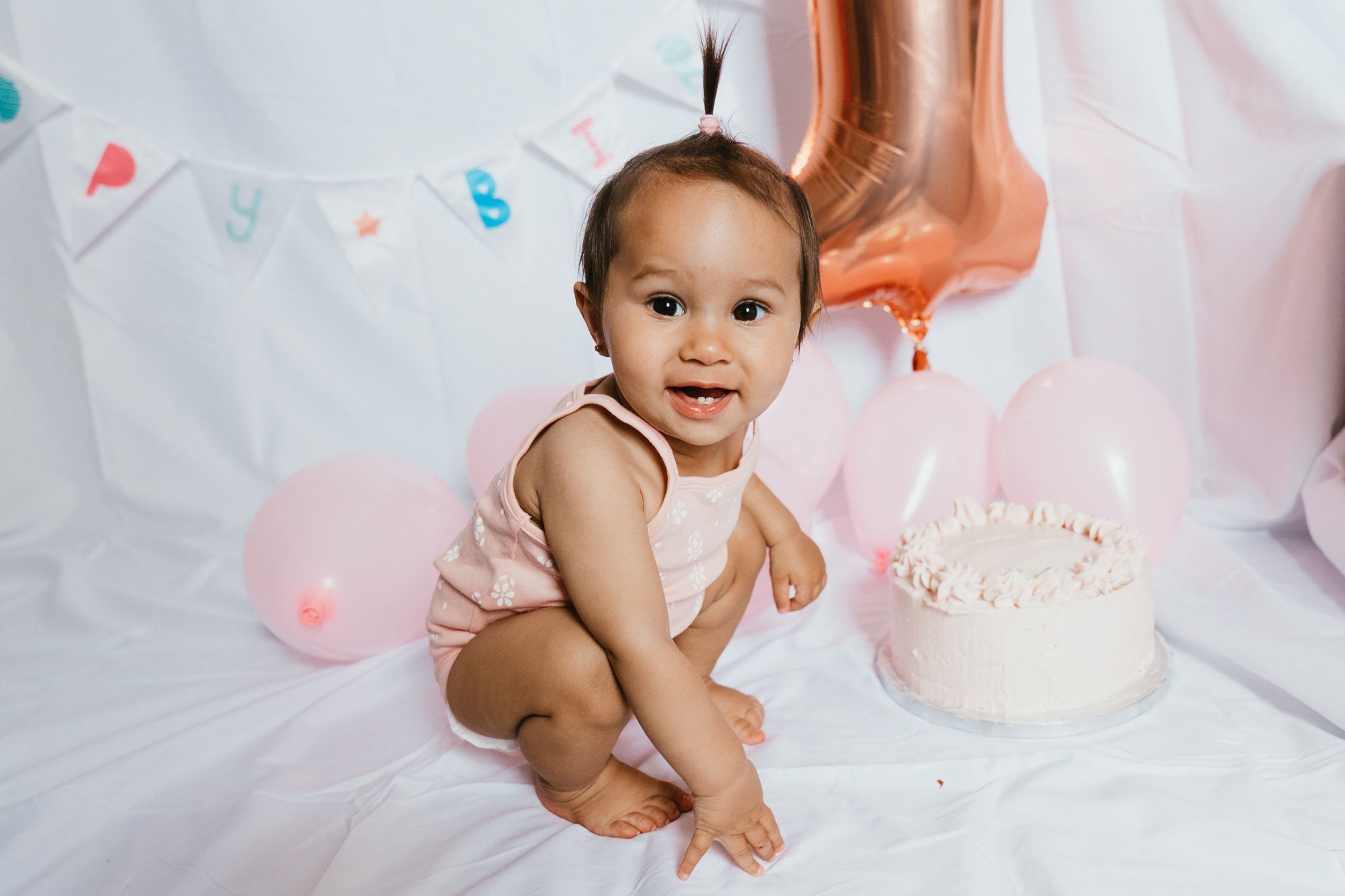 Baby in pink outfit sitting next to birthday cake with rose gold balloon decoration.