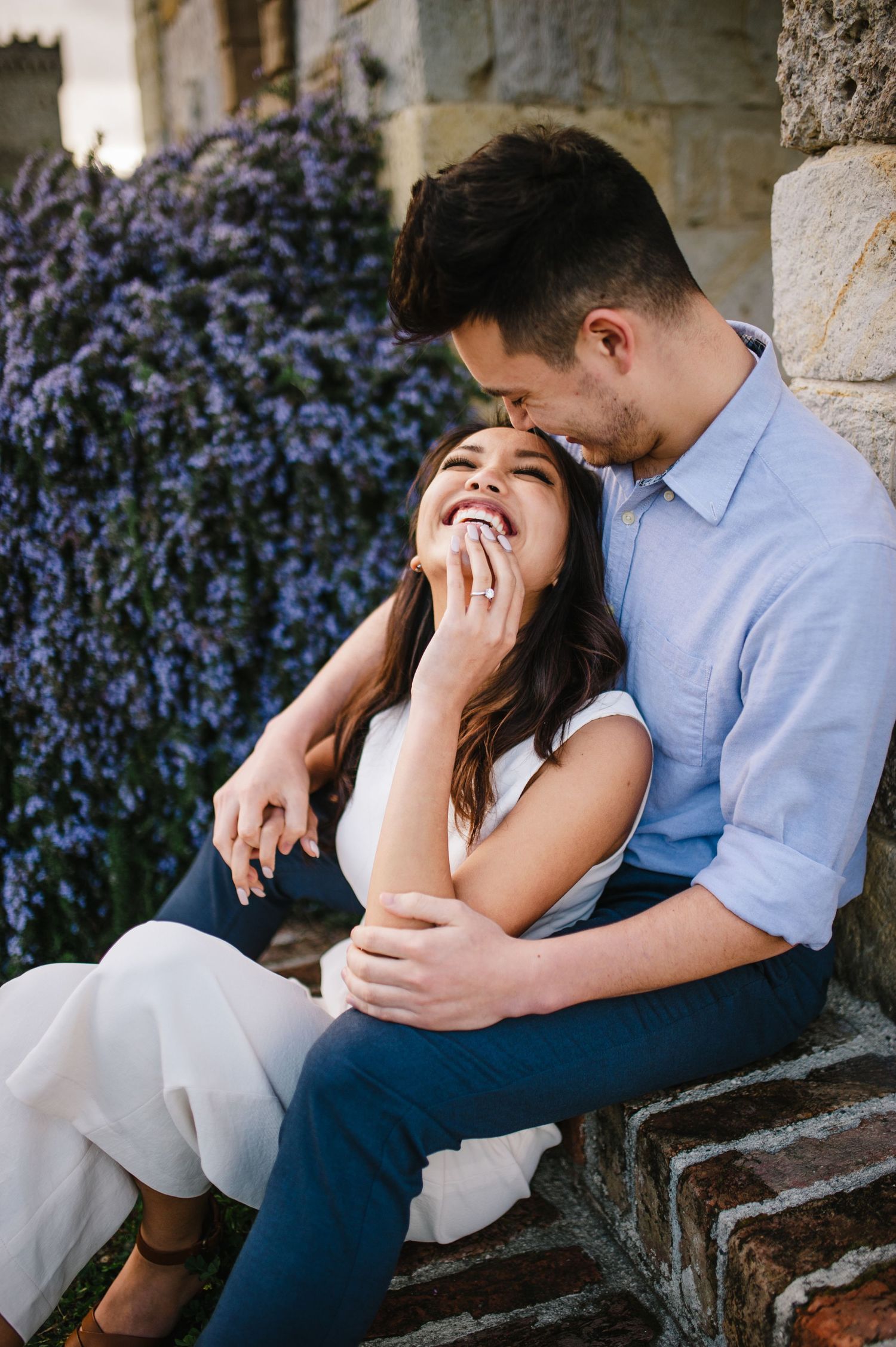 Candid moment of couple laughing together on stone steps with purple flowers in background.